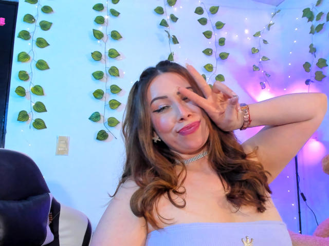 JennyLawry with wavy brown hair, wearing a strapless blue top, pink lipstick, choker, and watch, making a peace sign, smiling, showing her tits slightly. Bedroom with pink lights.