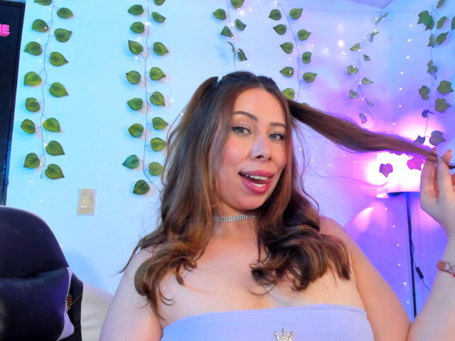 JennyLawry with wavy brown hair, holding a ponytail, wearing a strapless white top and a choker, smiling with pink lipstick, in a room with purple and blue lights.