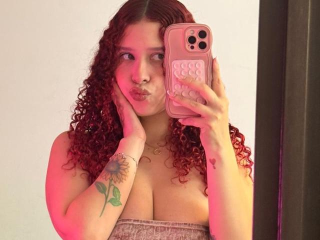 ValeryScot with curly red hair, pouty lips, holding a pink phone, wearing a strapless top, showing her tits, sunflower tattoo on right arm, pink and red lighting.