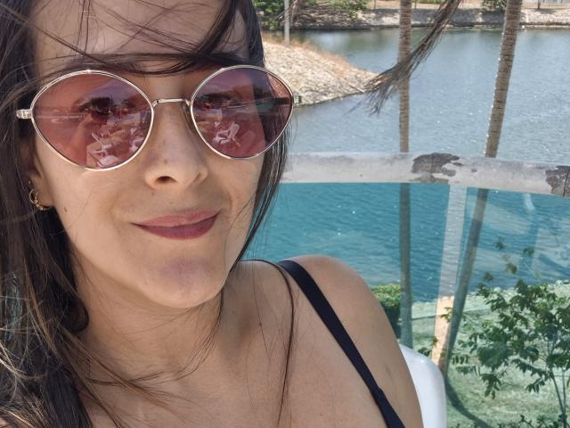 Roussemelinda69 porte des lunettes de soleil rondes avec une teinte rose, a les cheveux brun foncé et porte du rouge à lèvres. Elle porte un haut noir avec des bretelles qui dévoile sa poitrine, avec un fond blanc..