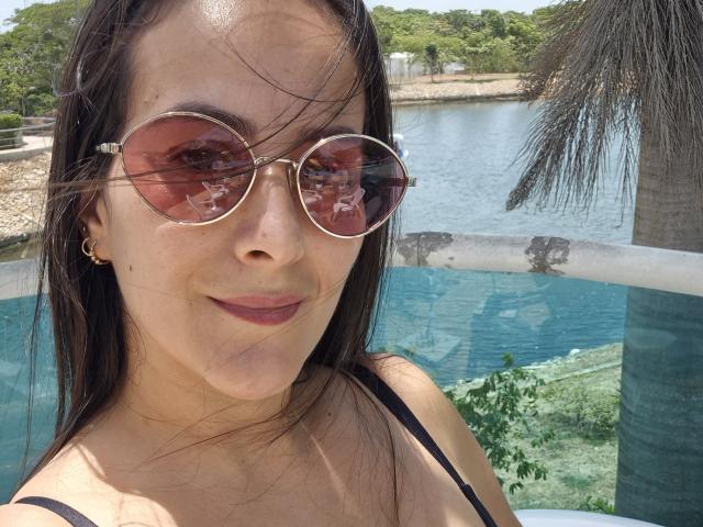 Roussemelinda69, avec des lunettes rondes teintées de rose, des cheveux bruns et un rouge à lèvres foncé, portant un haut noir à bretelles, souriant à la caméra, montrant ses seins et un petit piercing au nez..