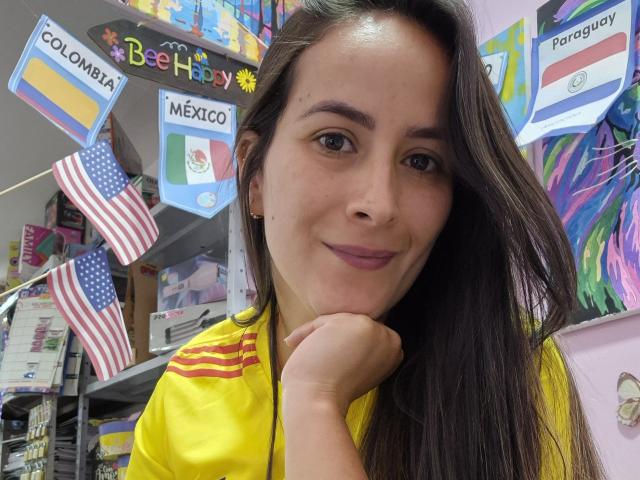 Roussemelinda69, com cabelos castanhos longos e batom roxo, apoia o queixo na mão, sorrindo, vestindo uma camisa amarela com listras vermelhas, em uma loja colorida..