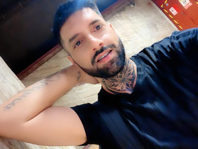 TommyDum, con barba, tatuaggi sul collo e sul braccio, maglietta nera e capelli ricci corti, sorride alla fotocamera, sdraiato su un pavimento di legno..