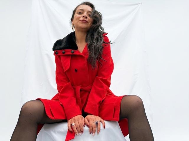 StangelarRivera, che indossa un cappotto rosso con colletto di pelliccia nera, collant neri e orecchini a cerchio dorati, è seduta con le gambe divaricate, sorridendo, con lunghi capelli grigi ondulati, e le mani appoggiate su una superficie bianca..