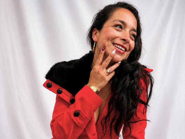 StangelarRivera con lunghi capelli neri, cappotto rosso con colletto di pelliccia nera, sorridente, mano vicino al viso, anello al dito, unghie rosse, sfondo bianco, che guarda di lato, sorridente..