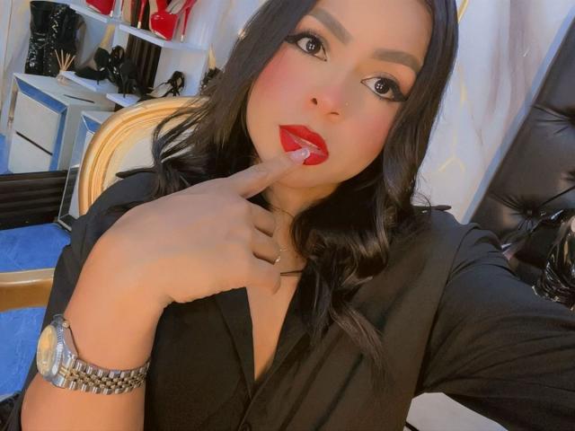 LindaFrancoo with red lipstick, black dress, and red finger in her mouth, wearing a silver watch, has long black hair and is looking at the camera.