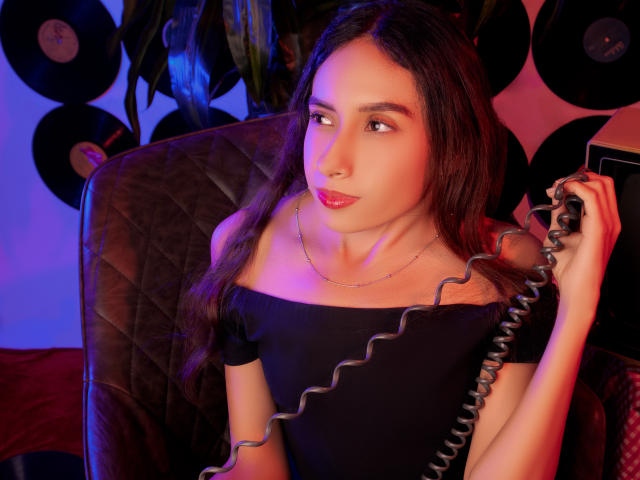 MiliBlom with long dark hair, off-shoulder black top, holding a coiled phone, sitting in a quilted chair, pink and blue lighting, looking to the side with red lipstick.