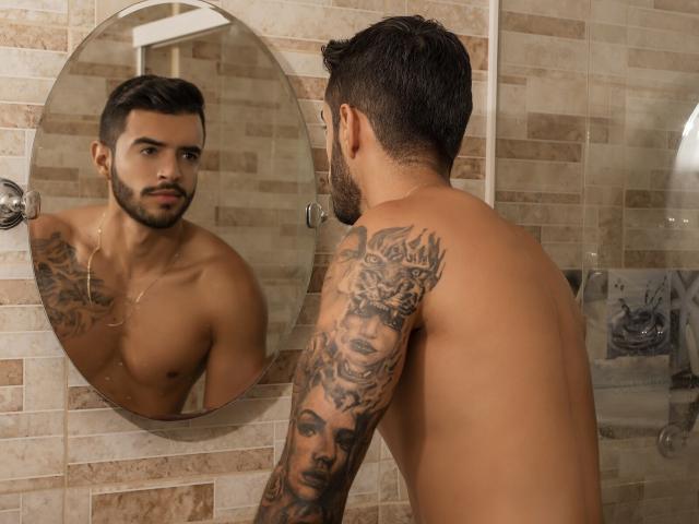 Shirtless Jessid with tattoos on right arm, standing in front of a mirror, looking at reflection, brown tile wall, short dark hair, beard, necklace, muscular build.