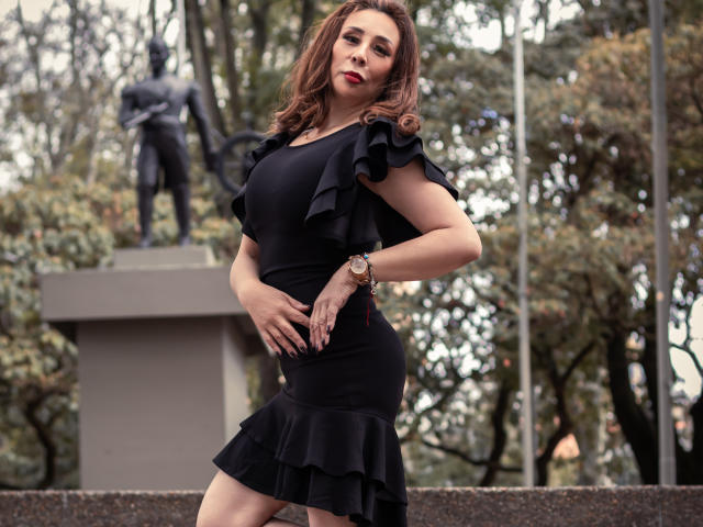 IssaMilfX with brown wavy hair, red lipstick, black ruffle dress, showing her tits, standing in front of a statue, hand on hip, wristwatch, confident, sexy.