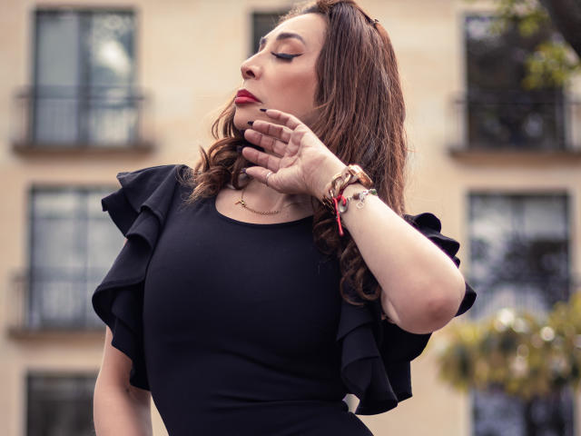 IssaMilfX with wavy brown hair, red lipstick, black ruffled dress, hand touching chin, gold bracelet, and red beads, standing outdoors, soft light, confident pose.