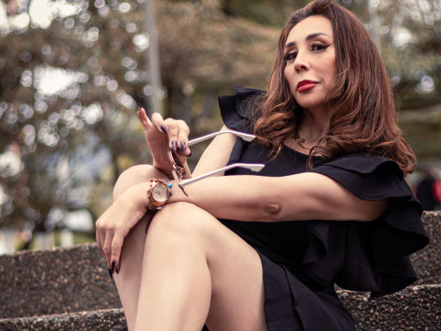 IssaMilfX with brown wavy hair, red lipstick, black dress, sitting with knees up, holding a pink ribbon, gold watch, and pink nail polish, looking off to the side.