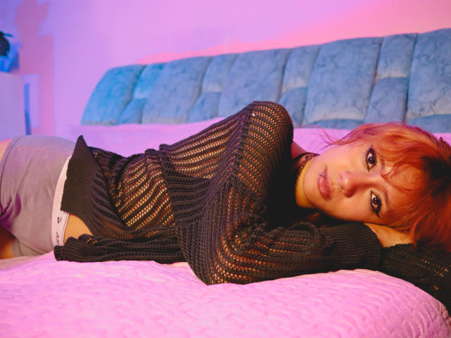 SuriEverly lies on a pink bed, wearing a black knit top, grey underwear, and a nose ring, with red hair and a sultry expression, in a purple-lit room.