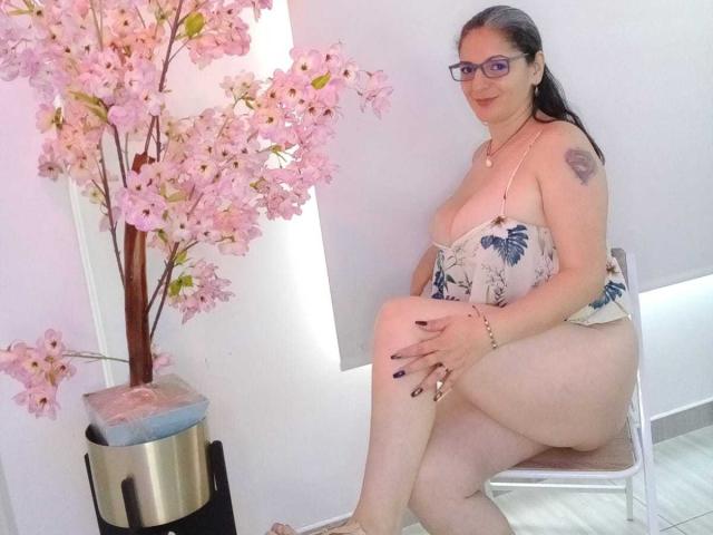 MadameScarlett, chubby with big tits, wearing a floral top, no pants, sitting on a chair, glasses, black nail polish, tattoo on shoulder, next to a pink cherry blossom plant.