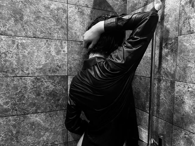 RubyMote with wet, dark hair, wearing a shiny black leather shirt, stands in a marble-tiled shower, hand covering her head, exposing her bare ass. Black and white photo.