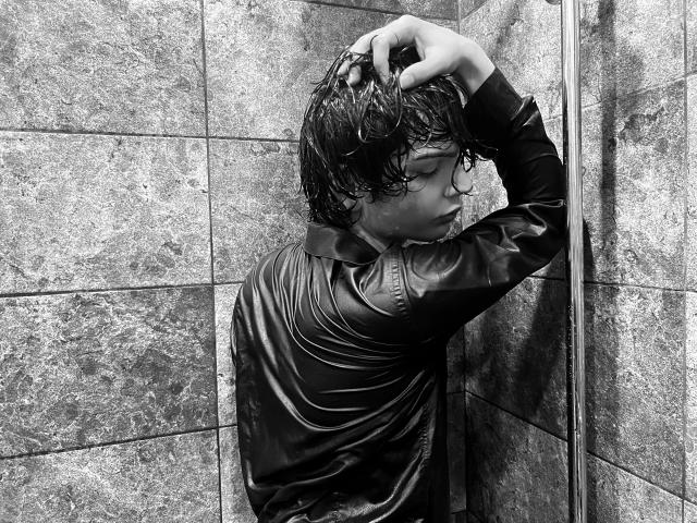 RubyMote with wet, dark hair, wearing a shiny black leather jacket, leans against a tiled wall, rubbing her head with one hand. Black-and-white, detailed, slightly moody.