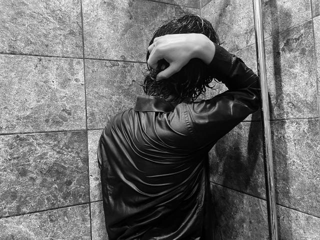 RubyMote, with wet, curly hair, wears a shiny black leather shirt, covering her head with one hand, in a tiled shower. Her back is to the camera.