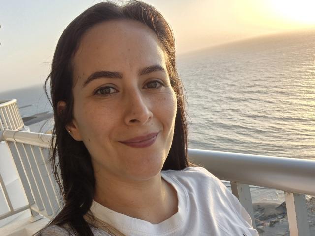 Roussemelinda69, with brown hair and light skin, smiling at the camera, wearing a white shirt, standing by a metal railing, ocean and sunset in the background.