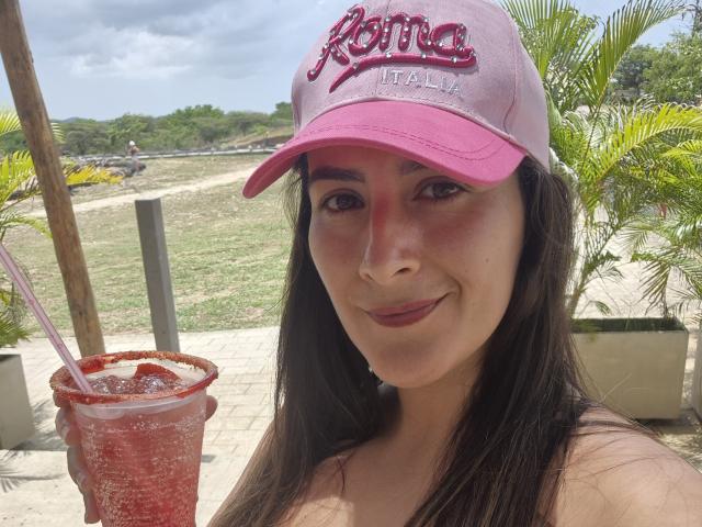 Roussemelinda69, avec le visage rouge, une casquette rose "Karma", de longs cheveux bruns, tenant une boisson rouge avec une paille, souriante, en extérieur, ensoleillé, portant du rouge à lèvres foncé, avec des marques de bronzage visibles, des palmiers en arrière-plan..