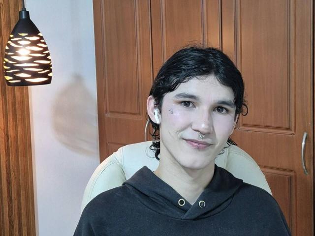Een vrouw met kort, nat zwart haar, een bruine hoodie, een neusring, een lip piercing en witte oordopjes, zittend in een beige stoel, lachend naar de camera..