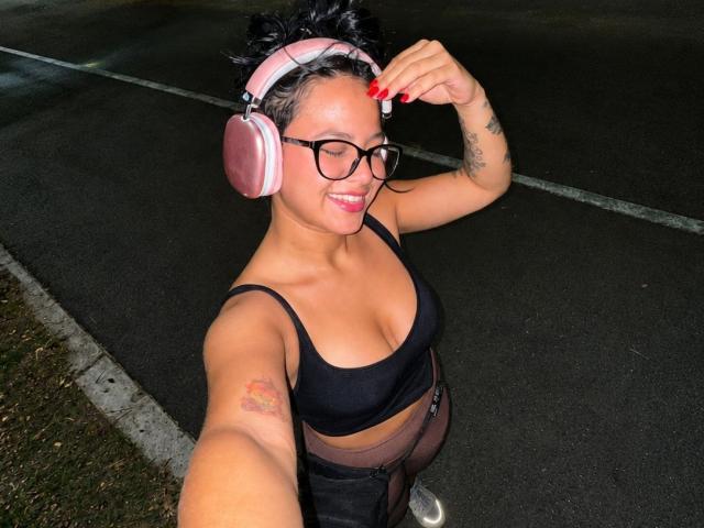 AlanaLuxe, com cabelo escuro em cachos, usando fones de ouvido rosa, sutiã preto e calças transparentes, sorrindo, com unhas vermelhas, tatuagem no braço superior, óculos, tirando uma selfie à noite..