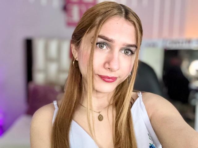 AndyDupont with long blonde hair, green eyes, pink lipstick, white top, small tits, necklace, earrings, and a slight smile in a selfie.