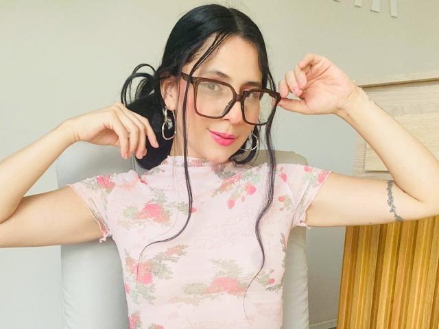 PeachSt with black hair, glasses, pink floral dress, holding glasses, smiling, pink lipstick, small tattoo on right arm, sitting in gray chair, wooden table in background.