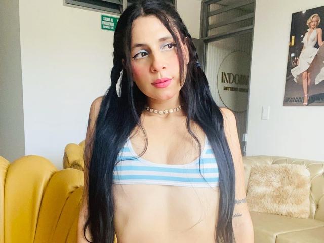 PeachSt, avec de longs cheveux noirs attachés en couettes, portant un soutien-gorge à rayures bleues, du blush rose, un collier ras du cou et des boucles d'oreilles cerceaux, se tient devant un canapé jaune. Un tatouage est visible sur le bras droit..