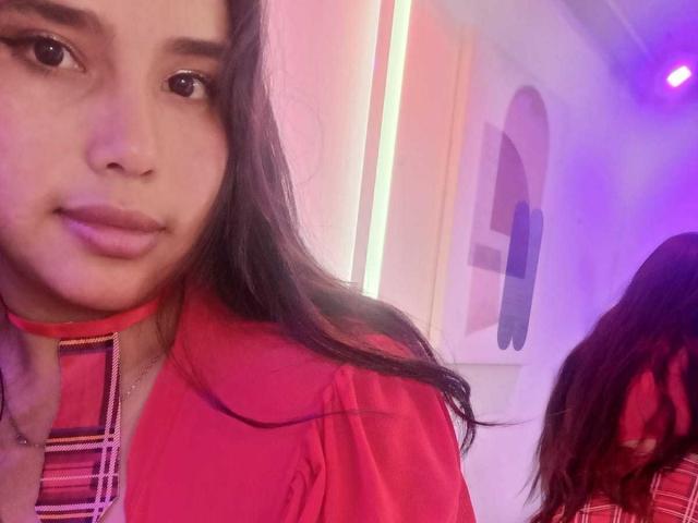 Gemma Bellini, com cabelos longos e pretos, camisa xadrez vermelha e colar, olhando para a câmera, luz rosa-roxo, sua bunda parcialmente visível no reflexo, sorriso suave, maquiagem discreta..