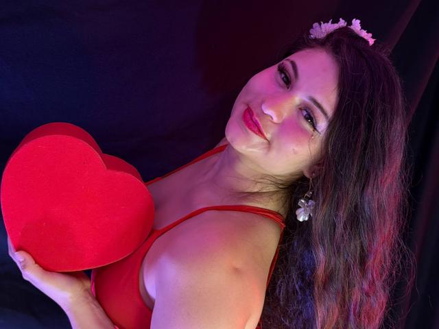 Esmaelda met lang bruin haar, rode lippenstift en een bloemenkrans, die een rood hart vasthoudt, een rode beha draagt en met een lichte glimlach naar de camera kijkt..