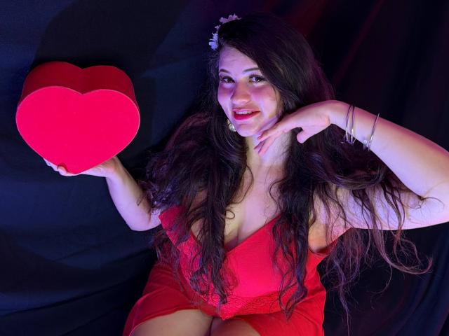 Esmaelda met lang donker haar, gekleed in een rode kantjurk, die een rood, hartvormig object vasthoudt, lachend, met een vinger op haar wang, op een zwarte achtergrond, met roze en paarse verlichting..