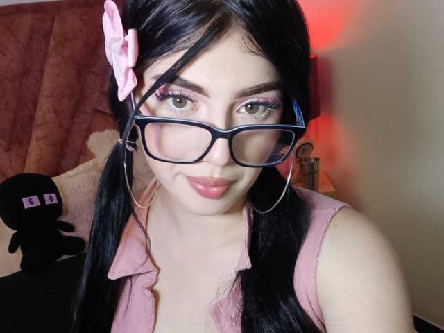 Isabella Rossi, con codini neri, fiocco rosa, occhiali, rossetto rosa, camicia rosa e orecchini a cerchio, mostra il seno, guardando la fotocamera con un leggero sorriso..