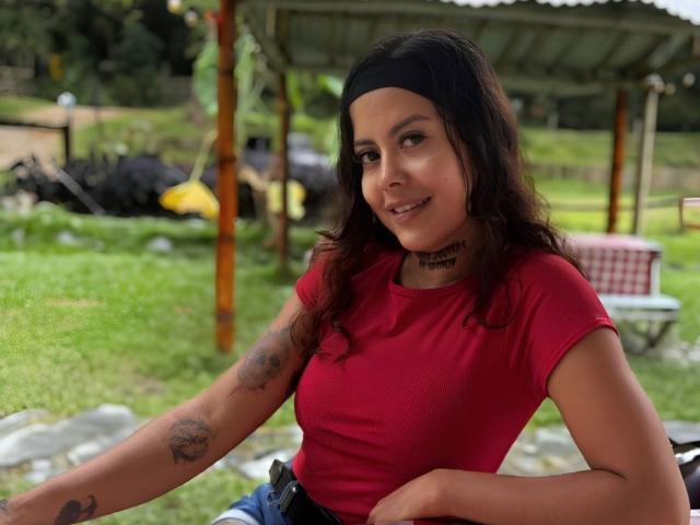 ElectraRoss met donker haar, een zwarte hoofdband, een rood shirt, tatoeages op de armen, lachend, met een gemiddelde borst, een tatoeage op de borst, een spijkerriem, zittend buiten, met groen gras, een houten constructie op de achtergrond..