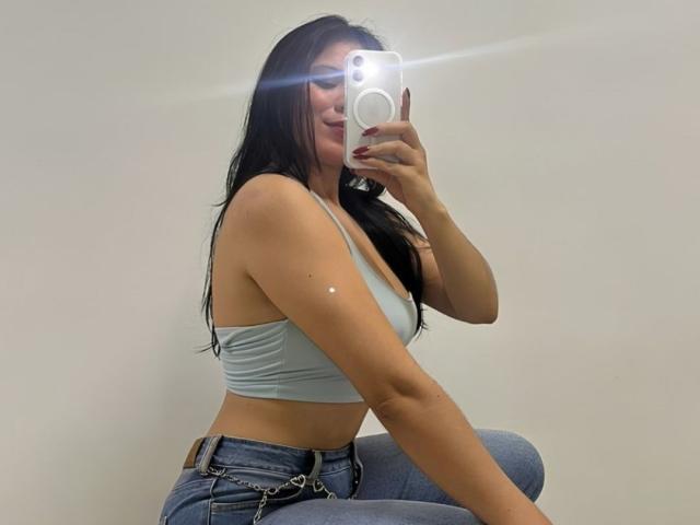 MaraCortes with long black hair, wearing a light gray sports bra and blue jeans, taking a mirror selfie, showing her small boobs and slim waist, smiling.