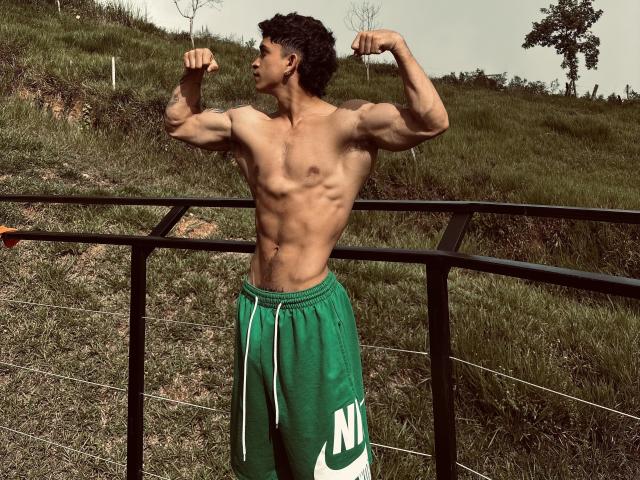 NoahhJacob, torse nu, contractant ses biceps, musclé, abdominaux bien définis, short Nike vert, debout à l'extérieur, sur une colline herbeuse, devant une clôture en métal noir, en journée, cheveux bruns bouclés, peau de couleur moyenne..