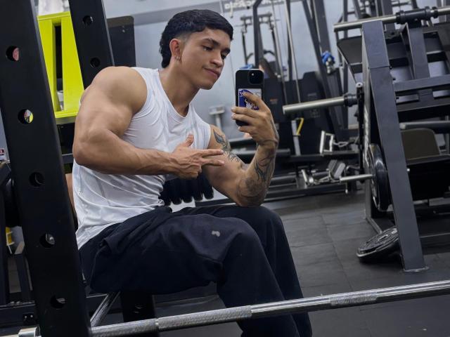 NoahhJacob, musclé, avec un bras tatoué, portant une chemise blanche sans manches et un pantalon noir, assis sur un banc de salle de sport, prenant un selfie avec sa main droite, souriant, sa main gauche pointant, dans une salle de sport moderne..