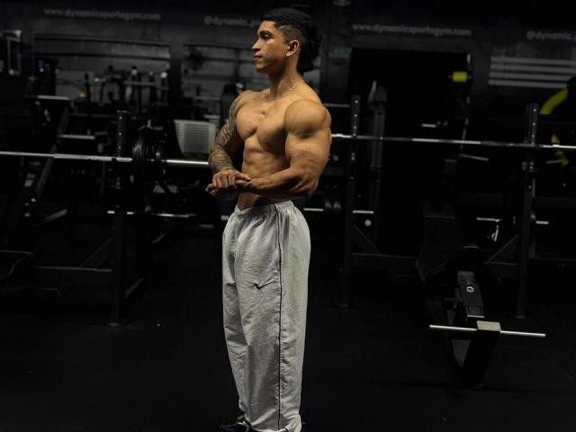 Muskulöser NoahhJacob mit tätowierten Armen, stehend in einem Fitnessstudio, trägt graue Nike-Hosen, Hände gefaltet, blickt zur Seite, dunkler Hintergrund, ernster Gesichtsausdruck, kurze, dunkle Haare, trainierter Hintern..