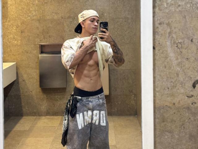 NoahhJacob, con un sedere tonico e muscoloso, alza la maglietta per mostrare i suoi addominali, indossa un cappello da baseball al contrario, pantaloni grigi con la scritta "MADE" e biancheria intima nera, e sta scattando un selfie allo specchio..