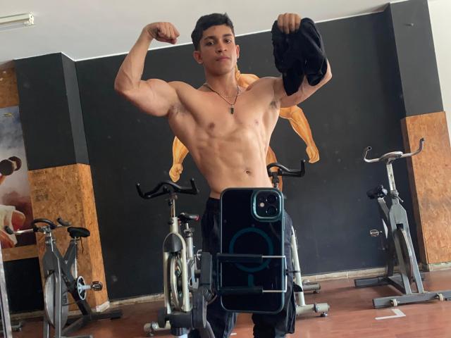 NoahhJacob, musclé, se montre dans une salle de sport, torse nu avec une serviette noire, qu'il tient dans sa main droite, portant un pantalon noir, debout devant des vélos d'exercice..