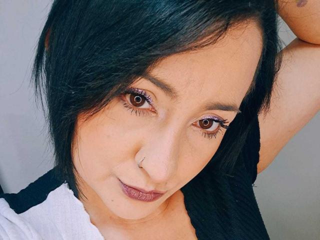 KiaRavoss has black hair, brown eyes, a nose ring, and a slight smile. She's wearing a black and white top with her arm raised.