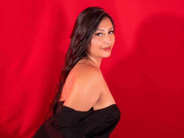 CatalinaRivero with long wet black hair, smiling, wearing a black off-shoulder top, exposing her tits, against a red background. She's looking over her shoulder.