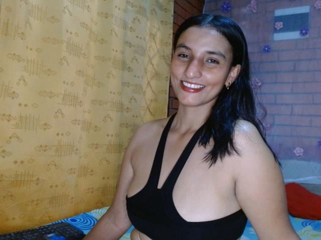 KeshaXsmith with dark hair, smiling, wearing a black sports bra that exposes her small, perky tits. She's standing in a room, looking happy and cute.