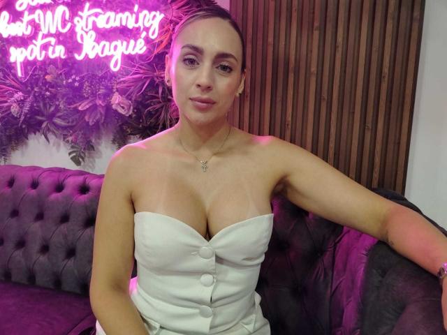 EvelynSwift, com seios pequenos, vestindo um vestido branco, sem alças e com botões, sentada em um sofá cinza, com detalhes em tufos, parede de madeira ao fundo, luz neon rosa acima, expressão calma, sorriso discreto..