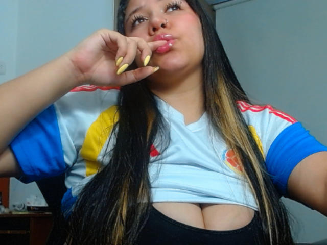 KrisGarner with long black hair, puffy pink lips, yellow nails, and a white shirt with blue and yellow sleeves, showing cleavage, pouting. Boobs partially visible under shirt.