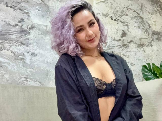 Elena Saenz, com cabelos cacheados roxos, sorrindo, vestindo um sutiã de renda preto e uma camisa preta aberta, expondo seus seios, em um quarto aconchegante com paredes semelhantes a mármore..