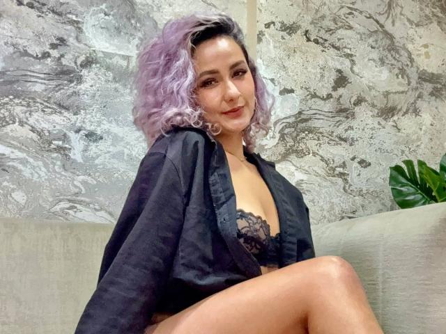 Elena Saenz, com cabelo cacheado roxo, sutiã de renda preto e camisa preta aberta, sentada em um sofá cinza, mostrando seus seios e sorrindo. Sua vulva não está visível..