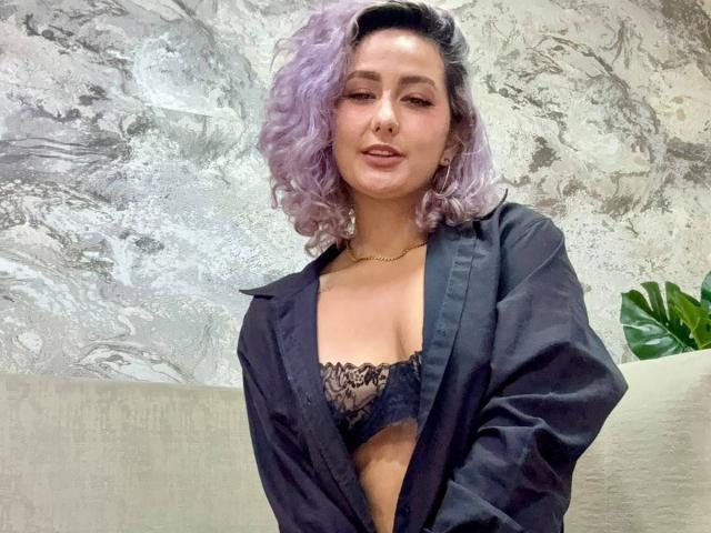Elena Saenz, com cabelos cacheados roxos, vestindo uma camisa preta aberta para mostrar seus seios, com um sutiã de renda, um colar de ouro e um leve sorriso..