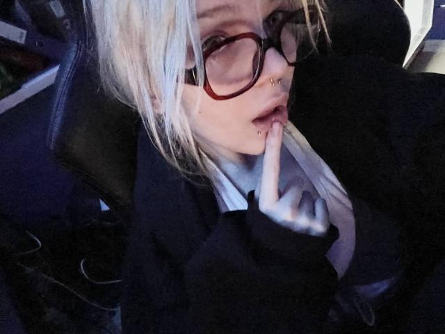 KinEvi, con cabello rubio, gafas, piercing en la nariz y en el labio, con un dedo en la boca, chaqueta negra, camisa blanca, mirando hacia arriba, dentro de un coche con poca luz..