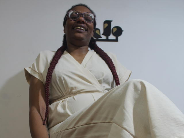 SimonaLujam with dark skin, glasses, and maroon braids, wearing a white dress, sitting with legs crossed, smiling, showing her tits slightly through the fabric.