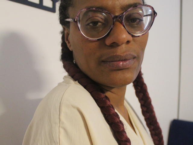 SimonaLujam with dark brown skin, braided red hair, and large purple glasses, wearing a cream shirt, has a serious expression, showing her small ass and cute nose.