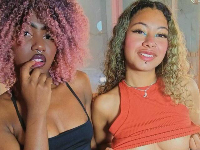 HaryAndKimmy with pink curly hair, nose ring, and finger in mouth, wearing black top. Kimmy with blonde curls, red top, and exposed tits, smiling. Both have pretty faces.